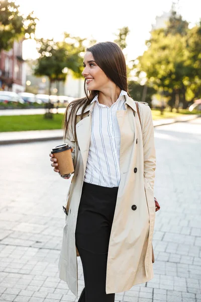 즐거운 귀여운 여자의 초상화 테이크 아웃 커피를 마시고 도시 거리에서 걷는 동안 옆으로 미소 — 스톡 사진