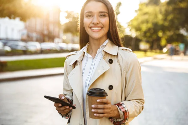 Portrét šťastné usmívající se ženy popíjí kávu a píšete si na mobil při chůzi po městské ulici — Stock fotografie