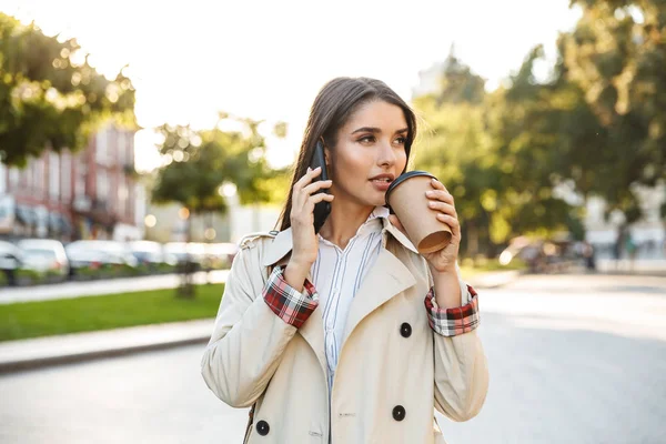 Portrait of thinking nice woman drinking takeaway coffee and talking on cellphone while walking on city street — Stock Photo, Image