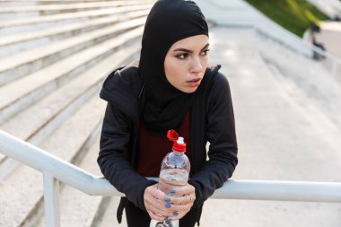 Genç ciddi müslüman spor fitness kadın başörtüsü ve koyu giysiler açık sokakta arka plan içme suyu tutan şişe tutan adımlar ile giyinmiş.