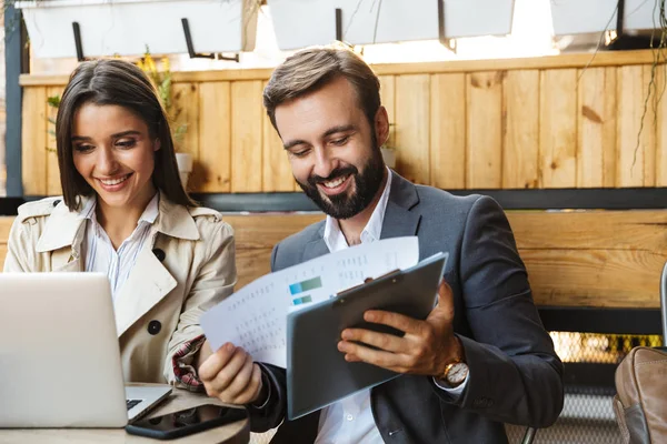 Foto de los trabajadores de oficina sonrientes hombre y mujer mirando los documentos y el ordenador portátil mientras usa el teléfono celular en la cafetería — Foto de Stock