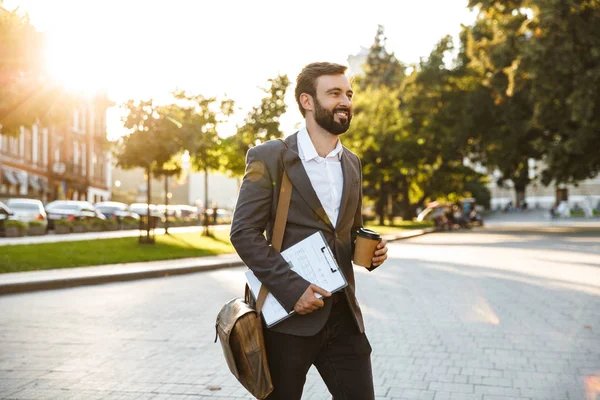Портрет красивого взрослого бизнесмена в официальном костюме, держащего кофе на вынос во время прогулки по городской улице — стоковое фото