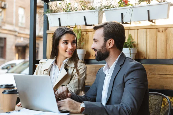 Retrato de belo casal de negócios homem e mulher conversando e trabalhando no laptop juntos enquanto sentados no café ao ar livre — Fotografia de Stock