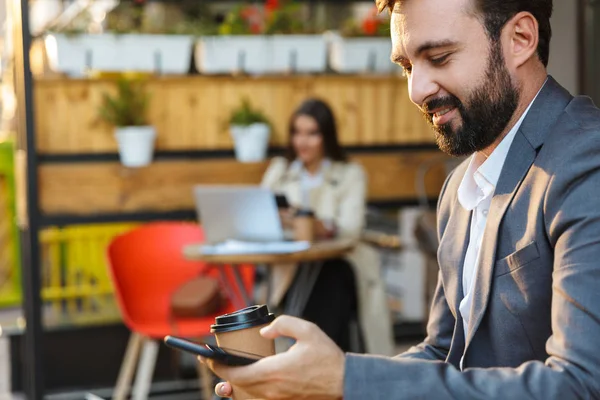 Portrait of smiling handsome man drinking takeaway coffee and typing on mobile phone while sitting in cafe — Stock Photo, Image