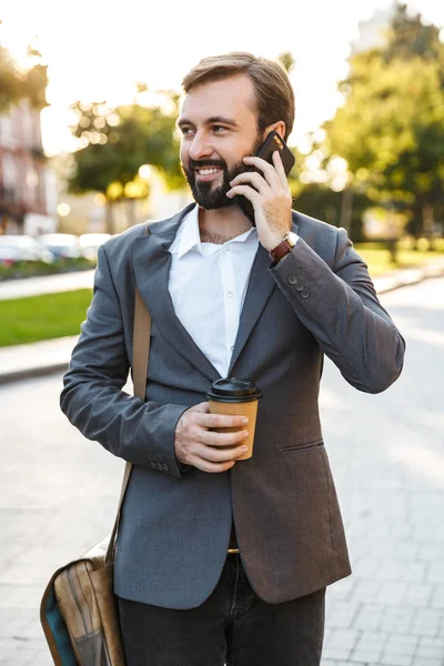 Porträt eines erwachsenen Geschäftsmannes im formellen Anzug, der im Freien Kaffee zum Mitnehmen hält und mit dem Smartphone spricht — Stockfoto