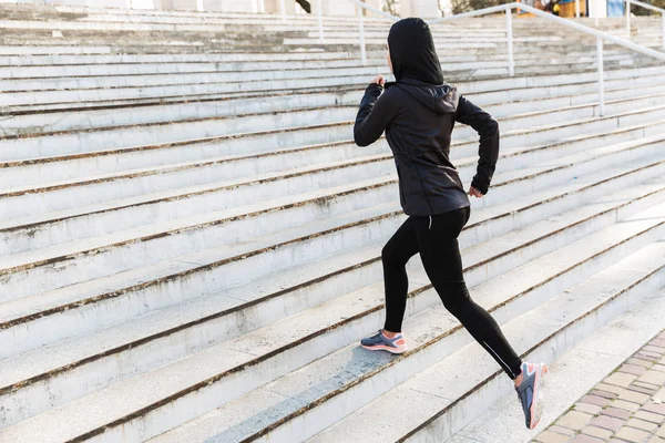 Young muslim sports fitness woman dressed in hijab and dark clothes running outdoors at the street.
