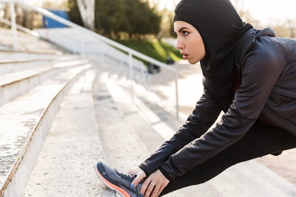 Muzułmańska Sport fitness kobieta ubrana w hidżab stwarzające zrobić sportowe ćwiczenia rozciągające na zewnątrz na ulicy z krokami na tle. — Zdjęcie stockowe