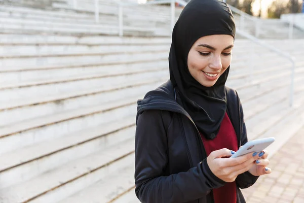 Wanita fitnes muslim yang ceria berpakaian jilbab dan pakaian gelap di luar rumah di jalan dengan langkah-langkah di latar belakang menggunakan telepon genggam . — Stok Foto