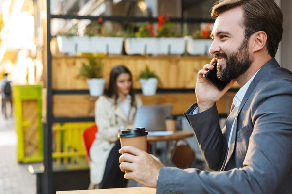 Retrato del hombre barbudo satisfecho bebiendo café para llevar y hablando por teléfono móvil mientras está sentado en la cafetería — Foto de Stock