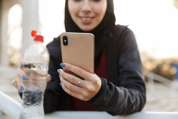 Junge konzentrierte muslimische Sport-Fitness-Frau in Hijab und dunkler Kleidung im Freien auf der Straße mit Schritten auf dem Hintergrund Trinkwasser haltende Flasche. — Stockfoto