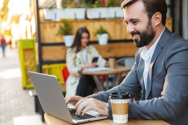 Portrait d'un bel homme d'affaires souriant et travaillant sur un ordinateur portable assis dans un café à l'extérieur — Photo