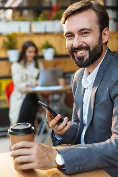 Портрет усміхненого щасливого чоловіка, який п'є каву і використовує мобільний телефон, сидячи в кафе — стокове фото