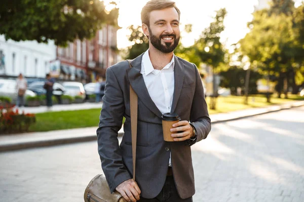 Portrait of bearded adult businessman in formal suit holding takeaway coffee while walking through city street — Stock Photo, Image