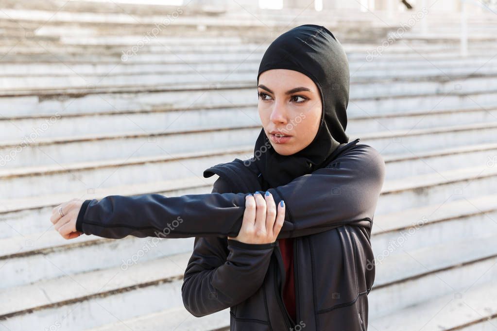Muslim serious sports fitness woman dressed in hijab posing make sport stretching exercises outdoors at the street with steps on background.