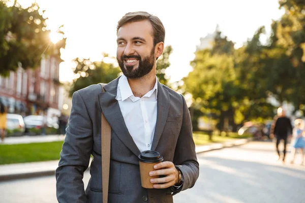 Portrait of smiling adult businessman in formal suit holding takeaway coffee while walking through city street — Stock Photo, Image