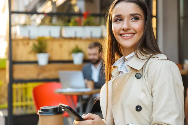 Πορτρέτο του χαμογελαστή ελκυστική γυναίκα πίνοντας καφέ σε πακέτο και πληκτρολογώντας στο κινητό τηλέφωνο ενώ κάθονται στο καφέ — Φωτογραφία Αρχείου