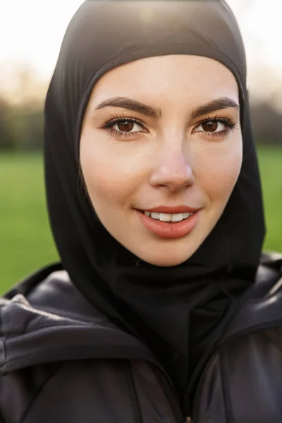 Retrato de primer plano de encantadora mujer musulmana atlética vestida con hiyab negro religioso sonriendo a la cámara en el parque verde al aire libre —  Fotos de Stock