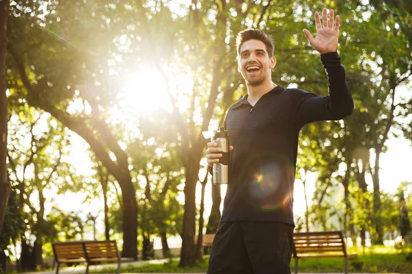 Jóképű vidám fiatal sport fitness ember állt a zöld Park természetvédelmi gazdaság palack vízzel integetett barátainak. — Stock Fotó