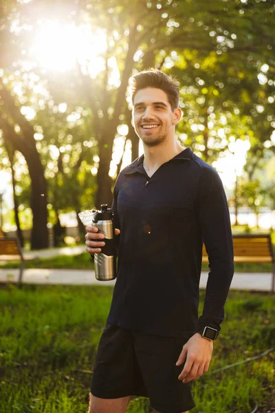 Jóképű mosolygó fiatal sport fitness ember állt a zöld Park természetvédelmi gazdaság palack vízzel. — Stock Fotó