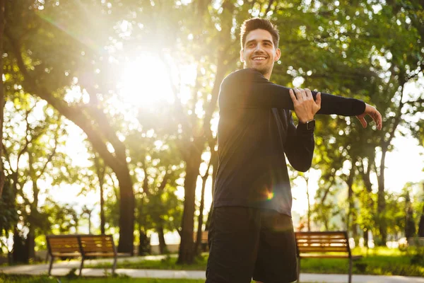 Cheerful young sports fitness man standing in green park nature make stretching exercises for arms. — Stock Photo, Image