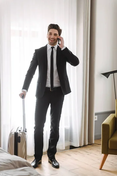 Foto av leende Europeiska affärsman talar på mobiltelefon och hålla bagage i Hotell Lägenhet — Stockfoto