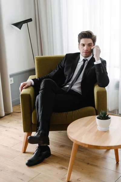 Photo of thinking handsome businessman talking on cellphone while sitting on armchair in hotel apartment — Stock Photo, Image