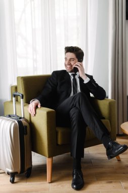 Otel dairesinde koltukta otururken cep telefonuyla konuşan gülen genç işadamının fotoğrafı