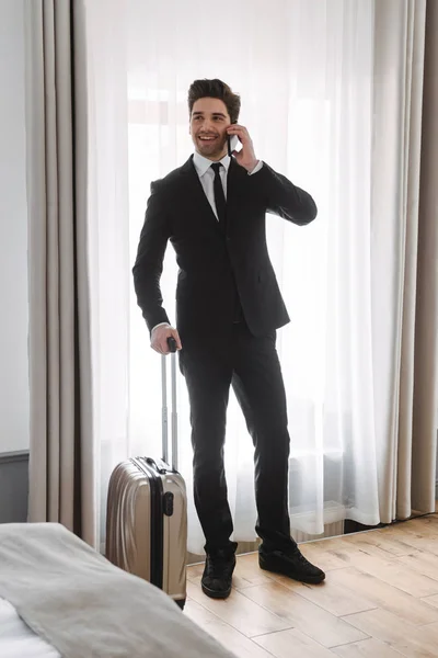 Foto de homem de negócios morena sorridente alking no celular e segurando bagagem no apartamento do hotel — Fotografia de Stock