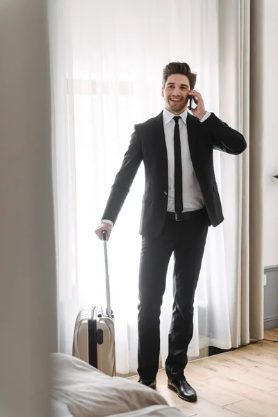 Foto av leende ung affärsman talar på mobiltelefon och hålla bagage i Hotell Lägenhet — Stockfoto