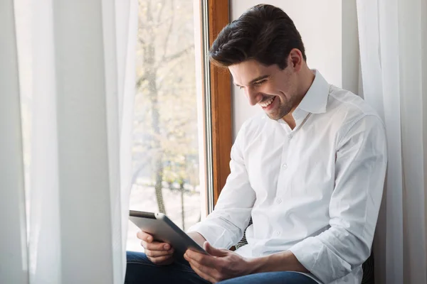 Photo of smiling unshaven man making video call on tablet computer while sitting at window in living room — Stock Photo, Image