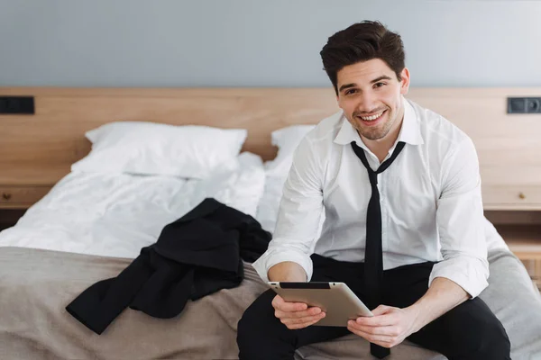 Photo of unshaven smiling businessman using tablet computer while sitting on bed in hotel apartment — Stock Photo, Image