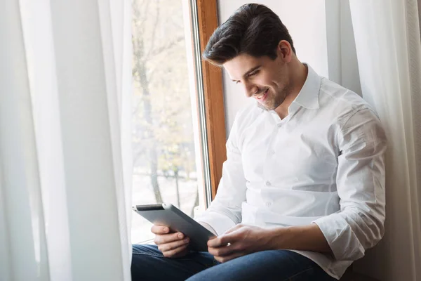 Photo of happy attractive man using tablet computer while sitting at window in living room — Stock Photo, Image
