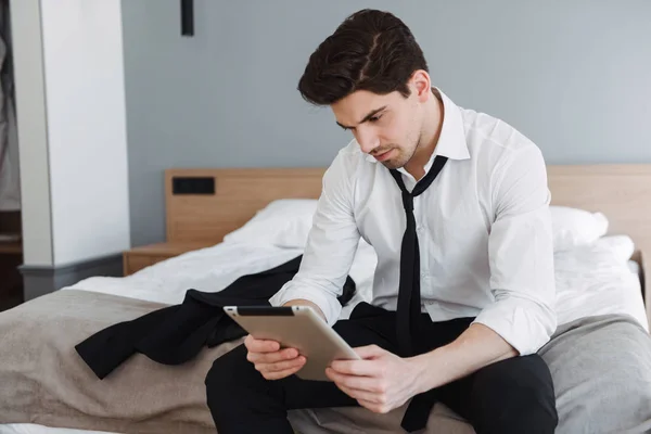 Photo of unshaven thinking businessman using tablet computer while sitting on bed in hotel apartment — Stock Photo, Image