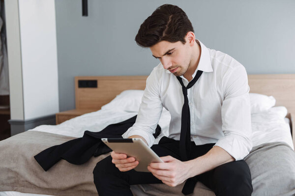 Photo of unshaven thinking businessman using tablet computer while sitting on bed in hotel apartment