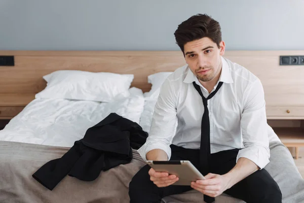 Foto de un hombre de negocios calmante sin afeitar usando una tableta mientras está sentado en la cama en el apartamento del hotel — Foto de Stock