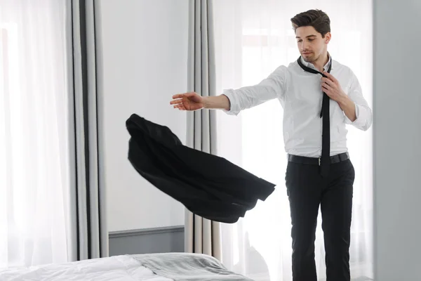 Täyspitkä kuva nuorten rauhoittava liikemies heittää takki sängylle hotellin huoneistossa — kuvapankkivalokuva