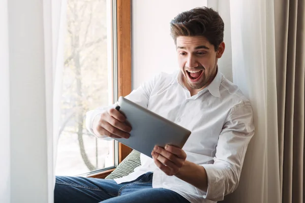 Photo of excited european man playing game on tablet computer while sitting at window in living room — Stock Photo, Image