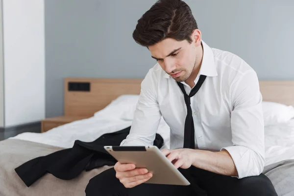 Foto de un hombre de negocios guapo pensando usando una tableta mientras está sentado en la cama en el apartamento del hotel —  Fotos de Stock