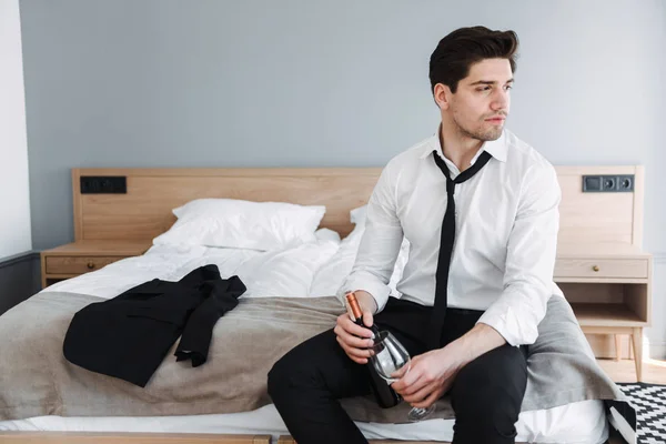 Foto de un hombre de negocios guapo morena sosteniendo una botella de vino y vidrio mientras está sentado en la cama en el apartamento del hotel —  Fotos de Stock