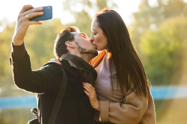 Felice giovane bella coppia amorevole posa passeggiando all'aperto nel parco natura prendere selfie da telefono cellulare baci . — Foto Stock