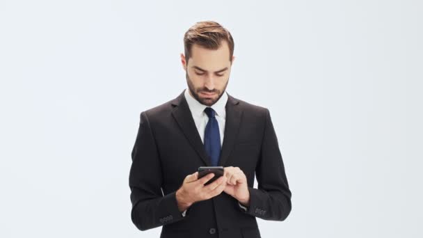 集中的年轻商人在黑色西装和蓝色领带变得惊讶和微笑 而使用智能手机在灰色的背景孤立 — 图库视频影像