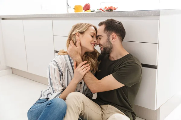 Feliz joven pareja cariñosa posando en la cocina en casa abrazándose besándose sentado en el suelo . — Foto de Stock