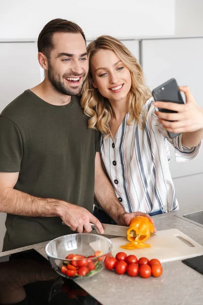 Jovem feliz casal amoroso posando na cozinha em casa abraçando tirar uma selfie . — Fotografia de Stock