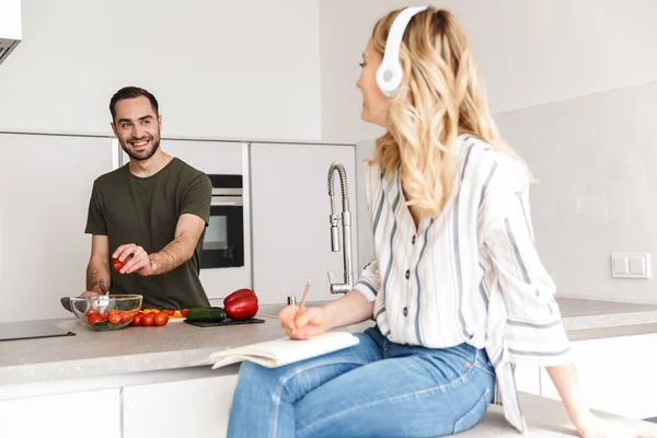 Jovem feliz dentro de casa na cozinha cozinhar salada de legumes perto de sua esposa ouvir música com fones de ouvido . — Fotografia de Stock