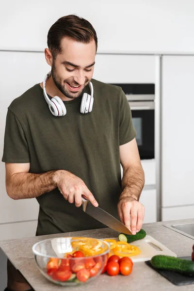 Jovem homem bonito feliz posando na cozinha em casa cozinhar ouvir música com fones de ouvido . — Fotografia de Stock