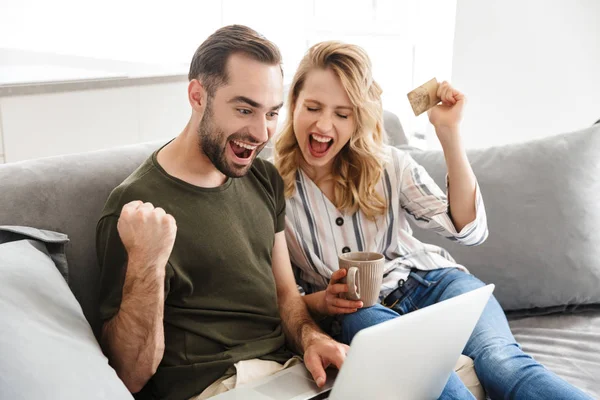 Szczęśliwy podekscytowany młody kochający para wewnątrz w Dom siedzący na sofa przy pomocy laptop rachmistrz zrobić zwycięzca gest posiadanie karta kredytowa. — Zdjęcie stockowe