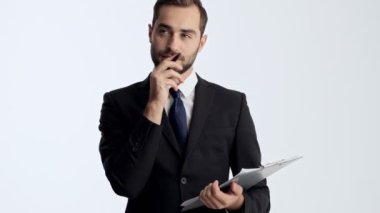Siyah takım elbise ve mavi kravat içinde pensive gülümseyen genç işadamı bir şey hakkında düşünme ve gri arka plan izole üzerinde belgelerde bazı notlar yapma