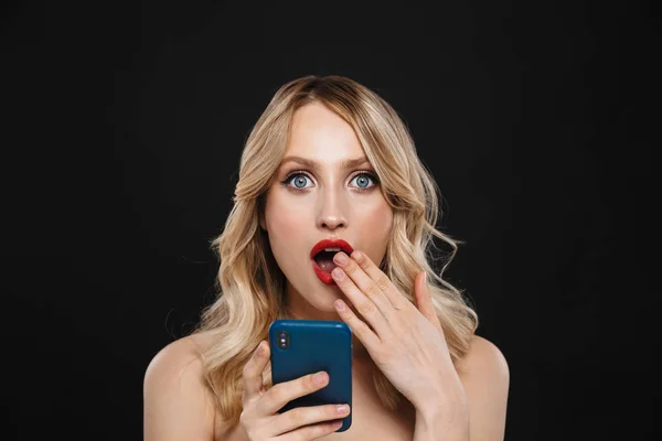 Шокована молода блондинка з яскравим макіяжем червоні губи позує ізольовано на фоні чорної стіни за допомогою мобільного телефону . — стокове фото