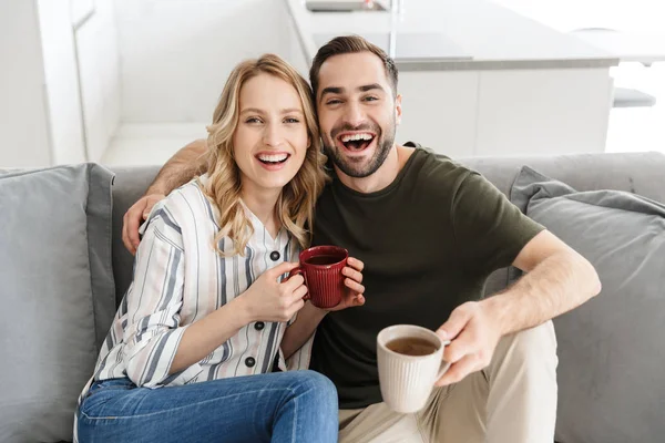 Feliz joven pareja amorosa sentada en el sofá en casa bebiendo té . — Foto de Stock