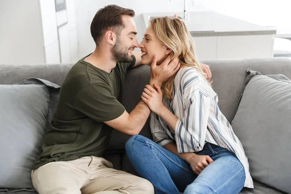 Smiling young loving couple indoors at home sitting on sofa hugging kissing. — Stock Photo, Image